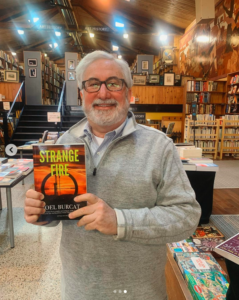 Joel Burcat is a white man with thick-rectangular glasses and quarter-zip sweater. He is standing in a book store and posing with his book.