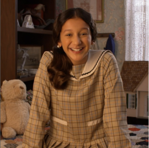 Dylan Tanzer is a 12-year-old white girl wearing a 1920s-style dress. She has long, brown hair brushed to one side of her shoulder and is smiling at the camera.