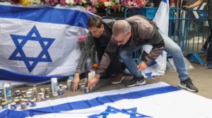 Two men with candles kneel on the ground in front of Israel flags hanging at a memorial.