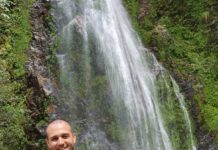 Tomer Morad is a white man wearing a bright red shirt and smiling at the camera. He is standing in front of a waterfall.