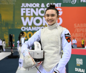 Maia Weintraub is wearing a full fencing uniform with her helmet tucked between her arm and foil in hand. Her dark hair is tied in a tight bun and she is smiling at the camera.