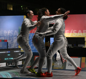 Four women in fencing uniform at embracing on a dimly lit stage.