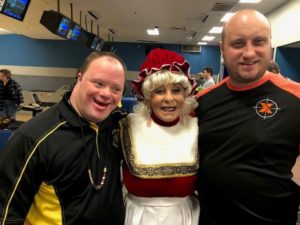 Elaine Brumberg is wearing a white wig and red hat. She is smiling and standing between two men.