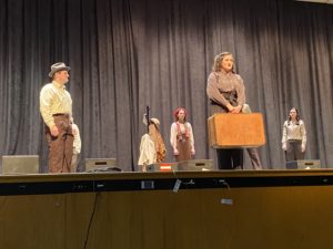 Standing on a stage are five actors in shabby 1940s-style clothing. One, center stage, is holding a suitcase.