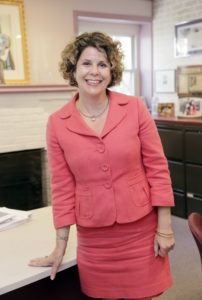 Leza Raffel is a white woman wearing a coral suit and skirt. She has shorter, curly brown hair.