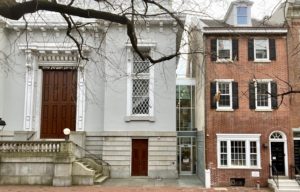 An old synagogue building painted a cool grey is connected by a glass door to a red brick Philly-style rowhouse.