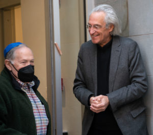 A short while man in a black mask and blue kippah is standing next to a taller older white man who is smiling at him. They are standing at the entrance of the Paula Kline Learning Center.