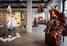 In an open arts space, abstract sculptures made of painted papier-mache and chicken wire are displayed on one side, while embroidered dresses and silver kitchenware are displayed on the other.