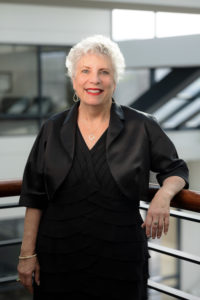 Becky Markowitz is a white woman with short, white hair and blue eyes. She is wearing a black dress and red lipstick, smiling.