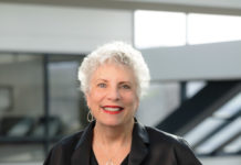 Becky Markowitz is a white woman with short, white hair and blue eyes. She is wearing a black dress and red lipstick, smiling.