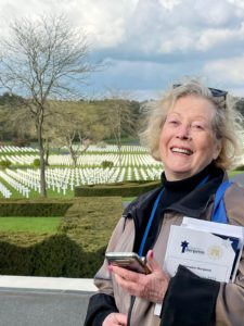 Barbara Belmont, a white woman with blonde-white hair, is smiling in front of a cemetery overlooking hundreds of graves.