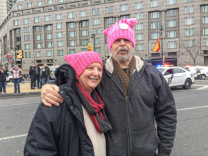 Standing outside in winter coats and pink "pussy hats" Carol Daniels, a white woman, and her husband, also white, stand with their arms around each other.