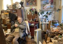 Philip Cohn is a white man sitting in a share in his studio, a small room filled with paintings, plants and knickknacks.