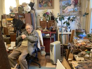 Philip Cohn is a white man sitting in a share in his studio, a small room filled with paintings, plants and knickknacks.