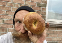 Philip Korshak is a white man with long beard and beanie. He is holding a bagel over one of his eyes.