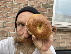 Philip Korshak is a white man with long beard and beanie. He is holding a bagel over one of his eyes.