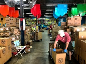 In a warehouse, volunteers pack boxes in a socially distanced manner.