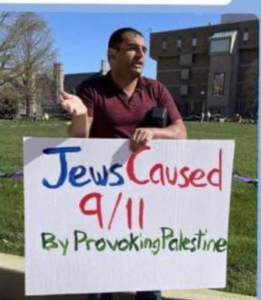 A man is standing outside holding a "Jews Caused 9/11 By Provoking Palestine" sign.