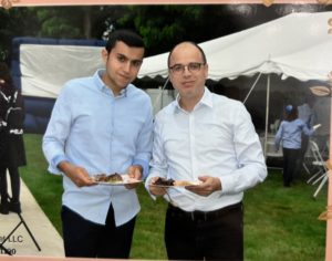 Moshe Asiag is an Israeli man wearing a blue button up shirt standing next to an older white man outside holding plates of food.