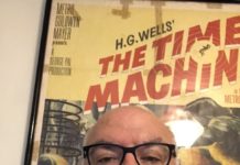 Irv Slifkin is a middle-aged white man who is bald and wear glasses. He is sitting in front of a movie poster.