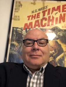 Irv Slifkin is a middle-aged white man who is bald and wear glasses. He is sitting in front of a movie poster.