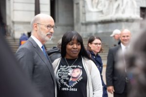 Tom Wolf is a white man with a thin white beard wearing a suit. He is standing next to a Black woman wearing a t-shirt of a deceased loved on.