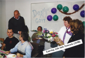 Several individuals sit around a conference room conversing and eating. A white board decorated with sparse balloons reads "DON'T GO"