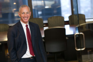 Ron Klasko is a middle-aged white man wearing a black suit and red tie. He is smiling and standing in front of a conference room.