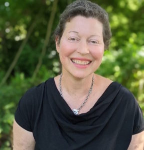 Gabrielle Kaplan-Mayer is a white woman with short, curly brown hair. She is wearing a black blouse and necklace and smiling at the camera outside.
