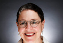 Debbie Albert is a white woman with short, black hair with a shock of white in it. She is wearing glasses and a white blouse, smiling in front of a black and white background.