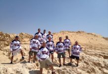 A group of men in red, white and blue hockey jerseys stand on a sandy desert mountain and smile for the camera.