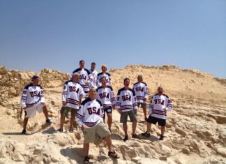 A group of men in red, white and blue hockey jerseys stand on a sandy desert mountain and smile for the camera.