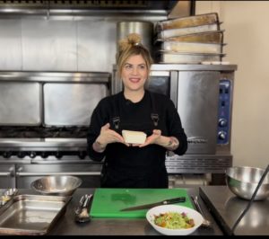 Rachel Klein is a white woman wearing a chef's coat hodling a block of tofu above a cutting board.