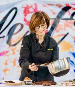 Fran Costigan is a white woman with red hair wearing a black chef's apron and holding a whisk and bowl filled with melted chocolate.