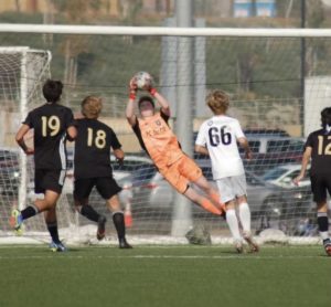 Jarett Wodotinsky is a white boy with red hair wearing a bright orange jersey. He is diving mid-air for a soccer ball as players on the other team approach the goal.