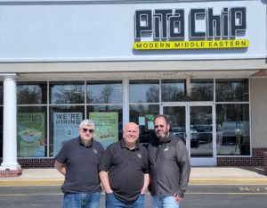 Standing in front of a storefront, three middle-aged men stand and smile.