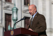 Doug Mastriano is a bald white man wearing a brown suit standing in front of a podium and microphone.