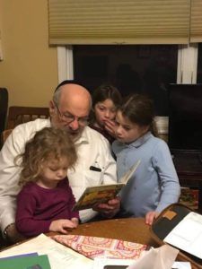 Gary Feldman is an old white man sitting on a chair crowded with three children as he reads a book.