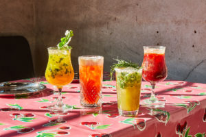 On a cherry-decorated table cloth, four fruity drinks sit in various glasses.