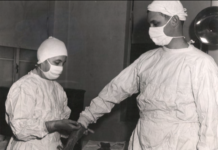 In a black and white photo, two doctors in full scrubs help each other don gloves for an operation.