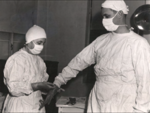 In a black and white photo, two doctors in full scrubs help each other don gloves for an operation.