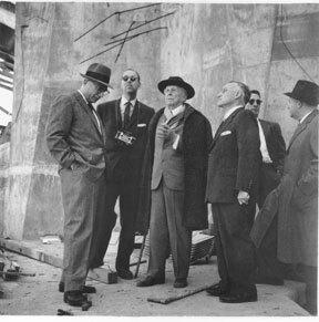 A black and white photos shows several men standing around in suits in conversation with each other.