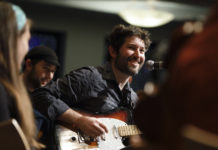 Joey Weisenberg is a white man with a scruffy beard and brown hair smiling as he plays the electric guitar.