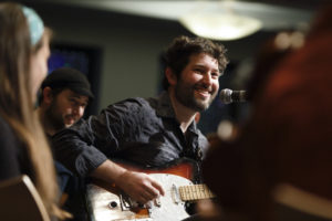 Joey Weisenberg is a white man with a scruffy beard and brown hair smiling as he plays the electric guitar.