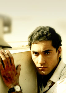 An Egyptian man with tidy hair and white shirt crouches on a balcony.