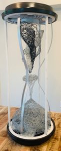 An hourglass made of recycled material has a spinning tornado on the top half, and an accumulating lump of chicken wire at the bottom.