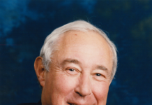 Frank G. Binswanger Jr. is pictured in the photo with a smile and a nice dress shirt, tie and suit jacket. The background is dark blue. His hair is white.