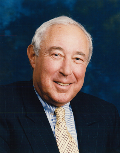 Frank G. Binswanger Jr. is pictured in the photo with a smile and a nice dress shirt, tie and suit jacket. The background is dark blue. His hair is white. 