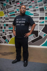 Jared Jackson is a black man with glasses and short, dark hair wearing a black t-shirt that reads "This is what JEWISH looks like."