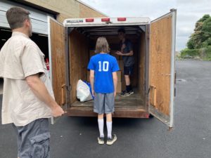 Standing in front of a trailer, a white man wearing a button up and shorts supervises two teenager loading up the trailer with boxes.